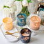 Decorative candles with floral designs on a marble surface with flowers and a mirror in the background.