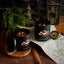 Two lit candles in black containers with 'Black Hills Candle' branding on a wooden surface with a map and compass.