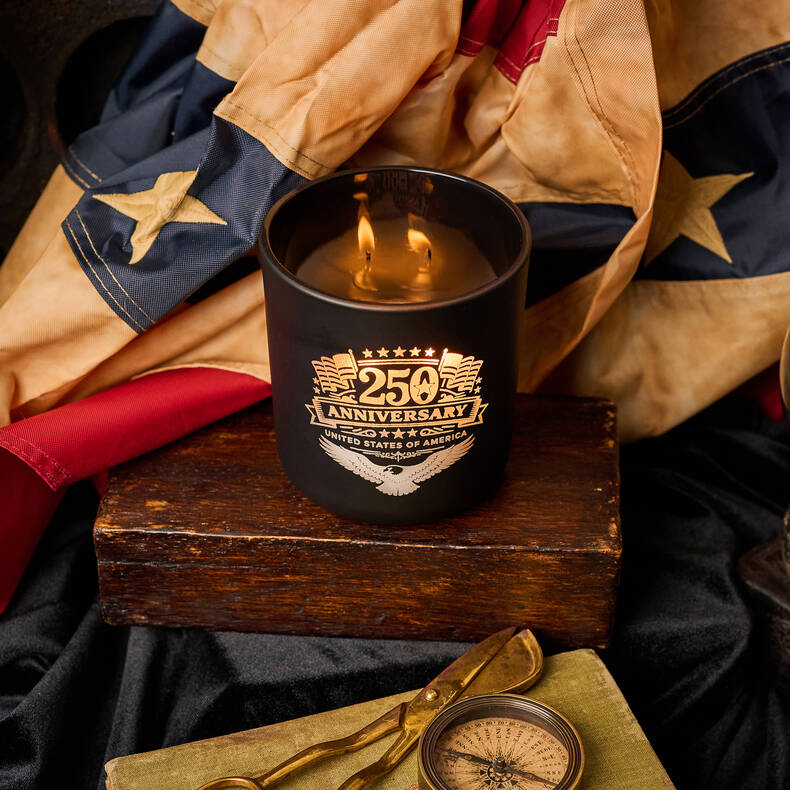 Black candle in a holder with '250th Anniversary' design on a wooden block, surrounded by a flag and compass.