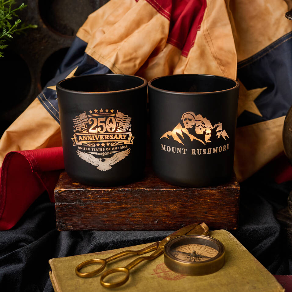 Two black mugs with gold text and graphics on a wooden box with vintage items.