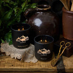 Two lit black candles with 'Black Hills Candle' branding on a rustic wooden surface with decorative elements.