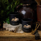 Two black candles with logo on a rustic wooden surface with decorative items. 