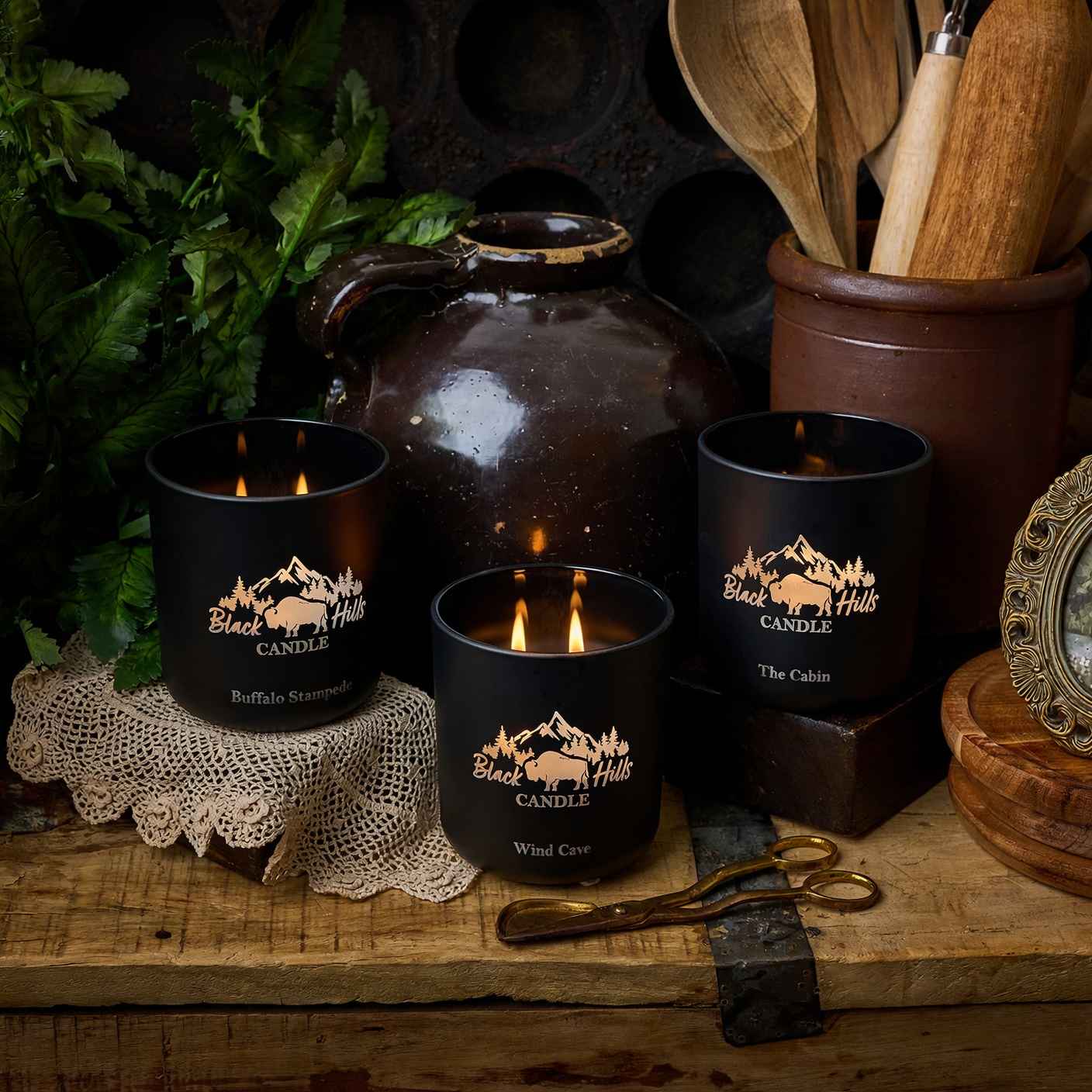 Black candles with 'Black Hills Candle' branding on a rustic wooden surface with a teapot and utensils. Main image