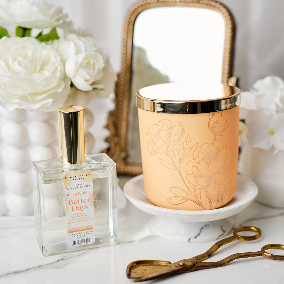 Perfume bottle labeled 'Better Days' next to a decorative peach cup with floral designs on a marble surface.
