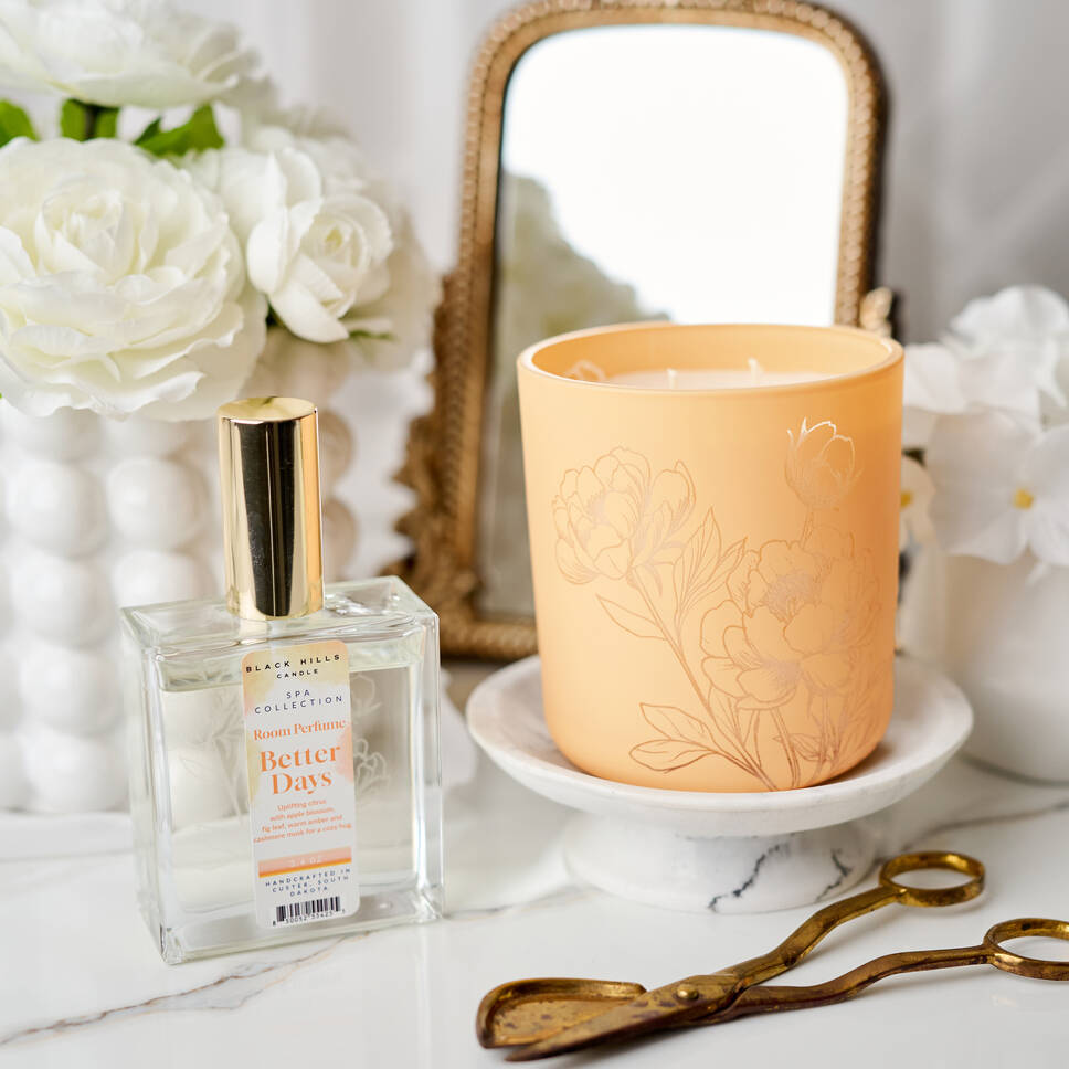 Perfume bottle and candle with floral design on a marble surface with flowers and mirror in the background.
