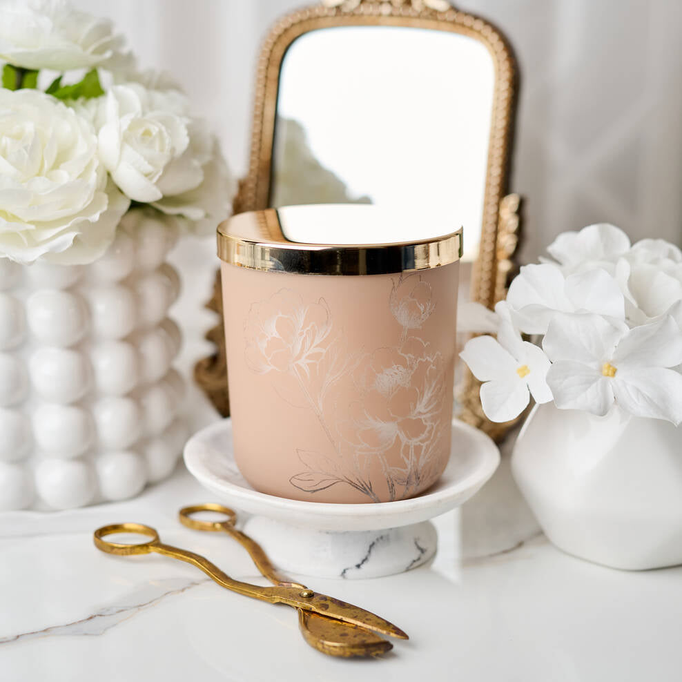 Decorative candle with gold lid on a marble surface with flowers and a mirror in the background