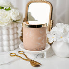 Decorative candle with gold lid on a marble surface with flowers and a mirror in the background