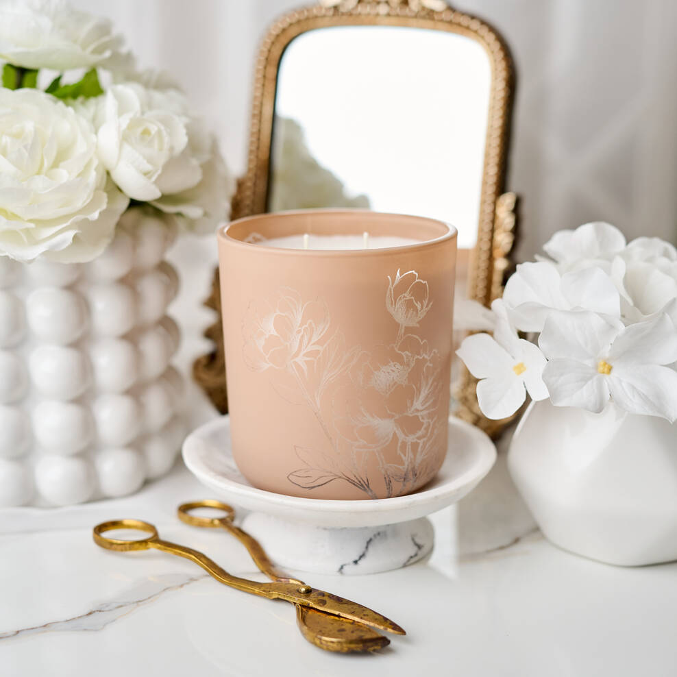 Candle with floral design on a white surface with flowers and a mirror in the background