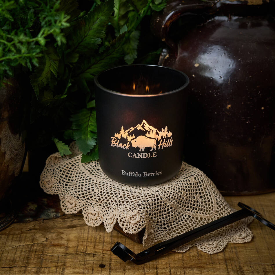 Black Hills Candle in a black container with a flame, placed on a lace doily with a rustic background. Main image