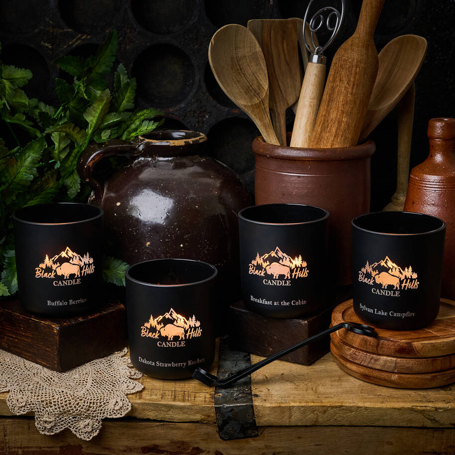 Black mugs with 'Black Hills Candle' logo on a rustic wooden surface with kitchen utensils and a jar.