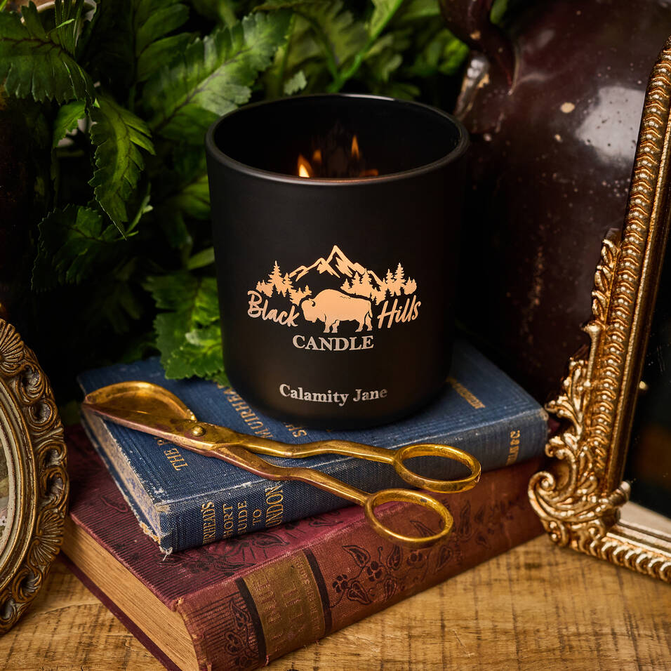 Black candle labeled 'Black Hills Candle Calamity Jane' on a stack of books with decorative items. Main image