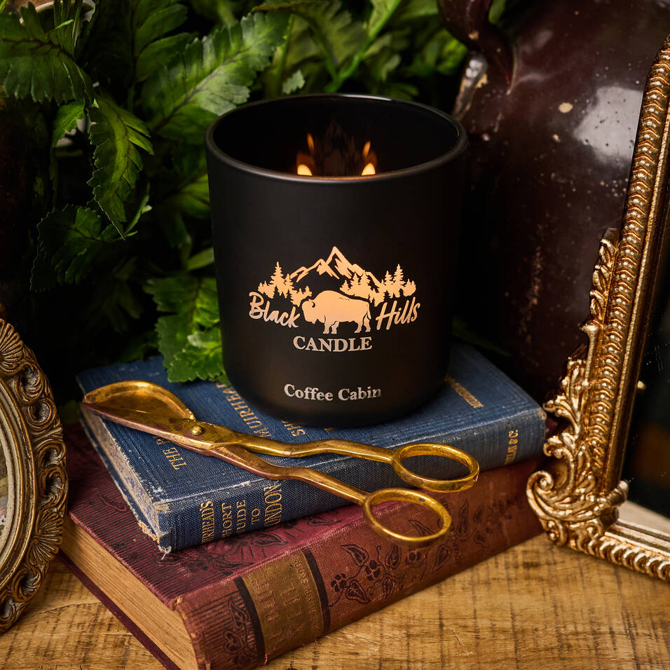 Black candle with 'Black Hills Candle Coffee Cabin' branding on a stack of books with gold scissors. Main image