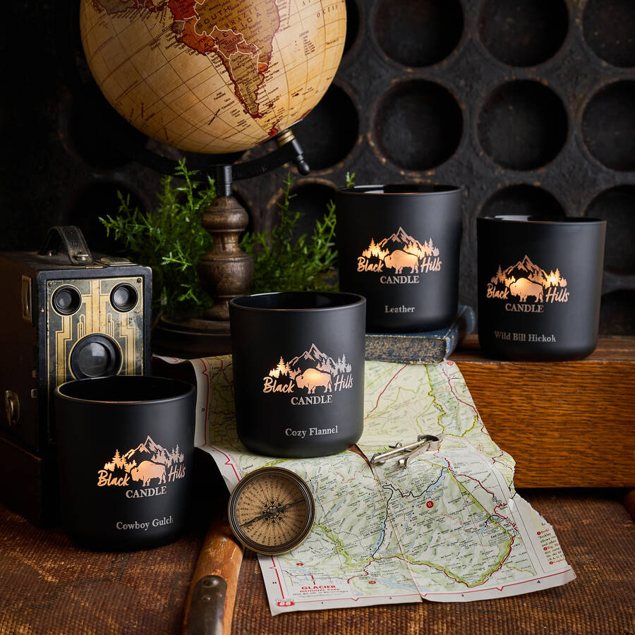 Black candles with mountain design and 'Black Hills Candle' branding on a rustic setting with a globe, map, and compass.
