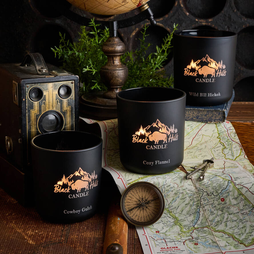 Black candles with 'Black Hills Candle' branding on a map with compass and vintage camera.
