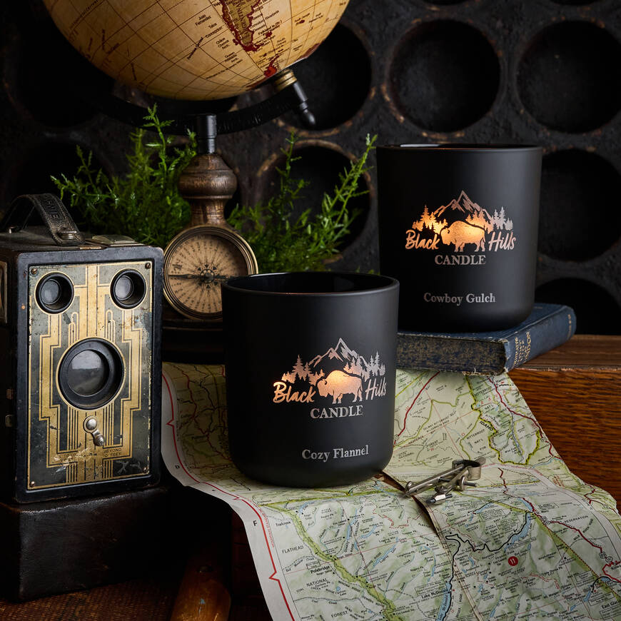 Two black candles with 'Black Hills Candle' branding on a map with vintage items.