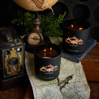 Two black candles with a logo on a map next to a vintage camera and globe.