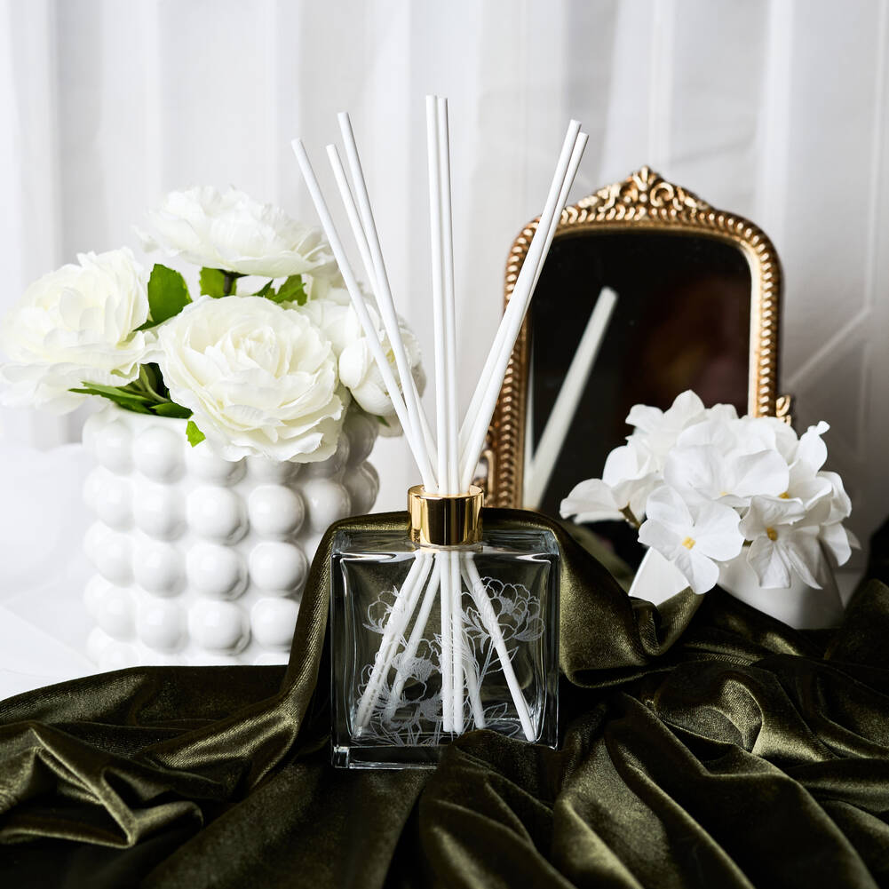 Diffuser with white sticks on a dark surface with flowers and a mirror in the background