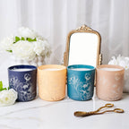 Four candles with floral designs on a marble surface with flowers and a mirror in the background.