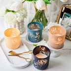 Decorative candles with floral designs on a marble surface with flowers and a mirror in the background.