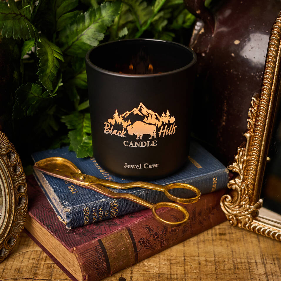Black candle with 'Black Hills Candle' logo on books with gold scissors and decorative frame.