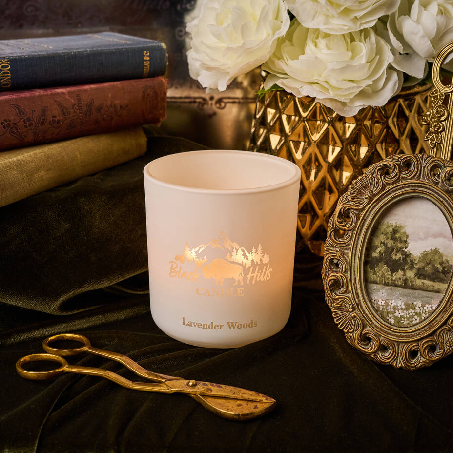Candle with 'Hills' logo on a dark surface with books, flowers, and a picture frame.