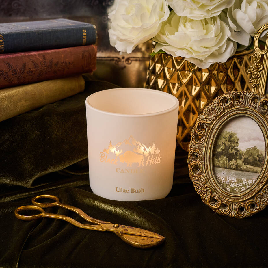 Candle with 'Canter Hills' branding on a dark surface with books, flowers, and a picture frame. Main image