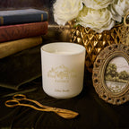 White candle with 'Black Hills Candle' branding on a decorative surface with books, flowers, and a picture frame.