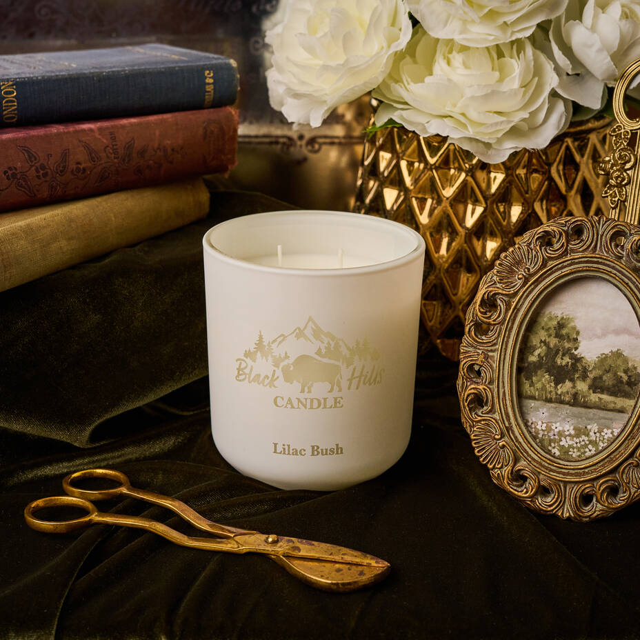 White candle with 'Black Hills Candle' branding on a decorative surface with books, flowers, and a picture frame.