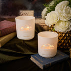 Two white candles with decorative labels on books, surrounded by flowers and books.