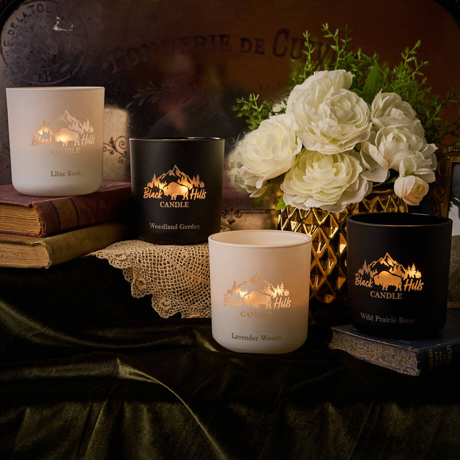 Collection of candles with decorative packaging on a dark surface with flowers and books. Main image