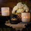 Three candles with decorative labels on a table with flowers and doilies.