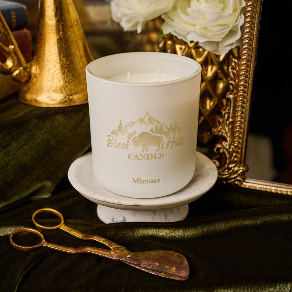 White candle with 'Black Hills Candle' branding on a decorative surface with gold accents and flowers.