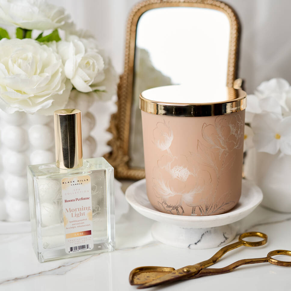 Perfume bottle and decorative candle on a marble surface with flowers and a mirror in the background.
