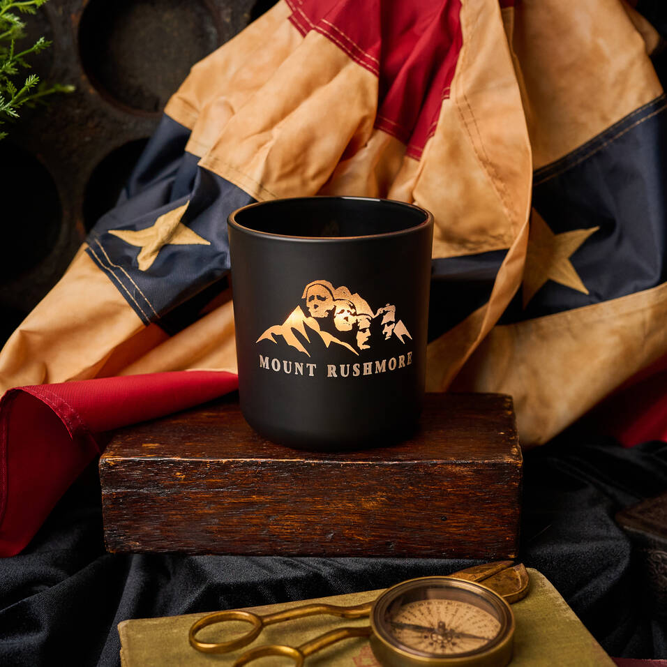 Black mug with 'Mount Rushmore' logo on a wooden block, surrounded by a jacket and compass.