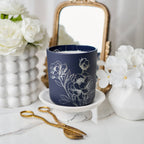 Navy blue candle with floral design on a white surface with flowers and a mirror in the background