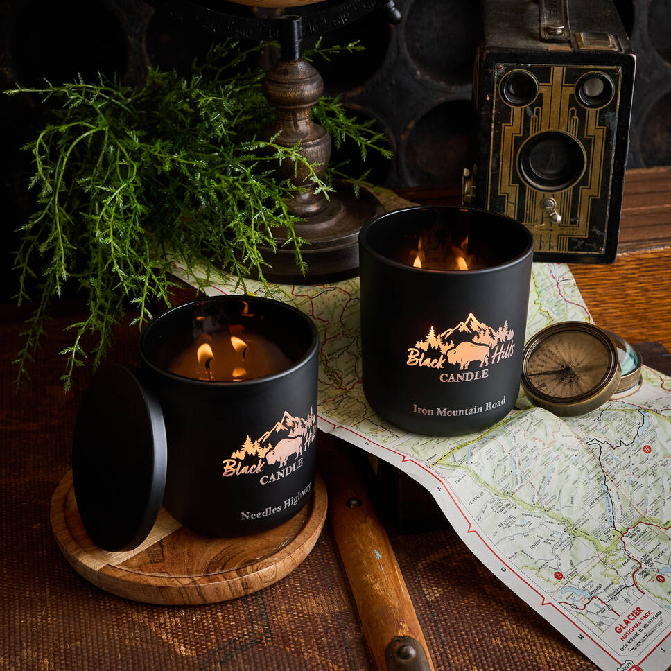 Two lit candles in black containers with 'Black Hills Candle' branding on a wooden surface with a map and compass. Main image