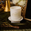 White candle with decorative design on a stand, surrounded by decorative items including a gold mirror and scissors.