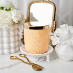 Rose gold candle with floral design on a white surface with flowers and a mirror in the background