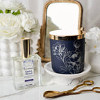Navy candle with floral design and perfume bottle on a white surface with flowers in the background