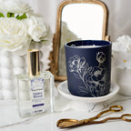 Navy candle with floral design on a white surface next to a perfume bottle and gold tongs.