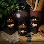 Three lit candles in black containers with a rustic setting featuring a wooden pot and kitchen utensils.