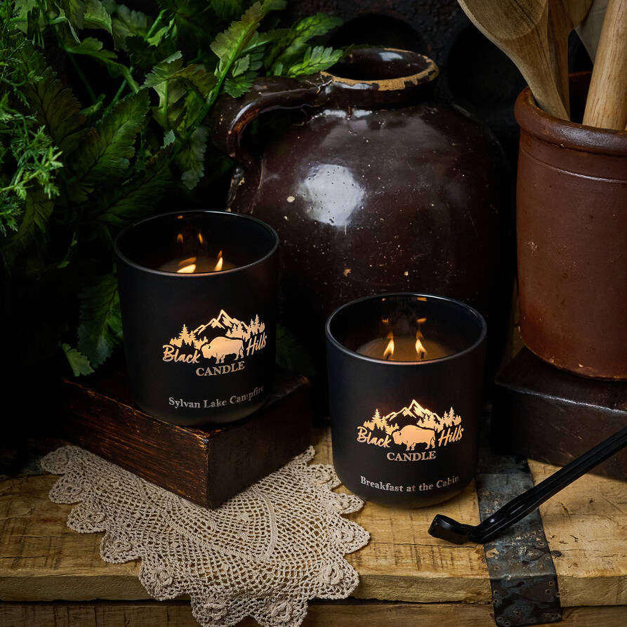 Two lit candles in black holders with a mountain logo on a rustic wooden surface.