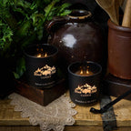 Two lit candles in black holders with a mountain logo on a rustic wooden surface.