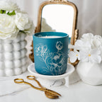 Blue candle with floral design on a white surface with flowers and a mirror in the background