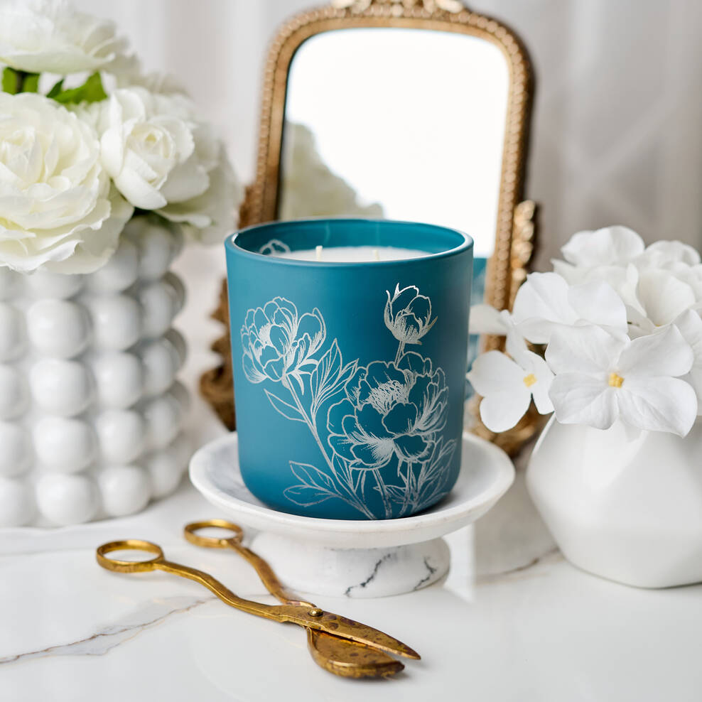 Blue candle with floral design on a white surface with flowers and a mirror in the background