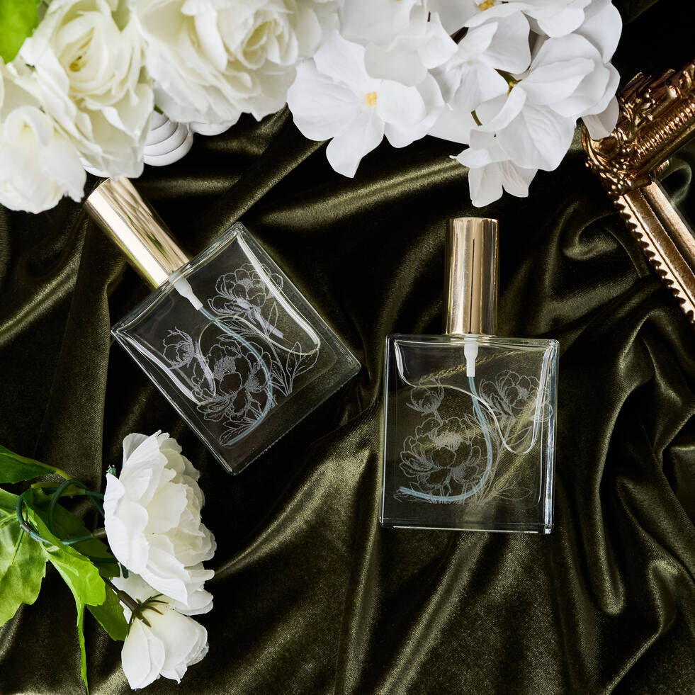 Two elegant perfume bottles with floral designs on a dark fabric background with white flowers.