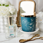 Perfume bottle and blue candle with floral design on a decorative surface with flowers and a mirror in the background.