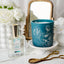 Teal candle with floral design and perfume bottle on a marble surface with flowers and a mirror in the background.
