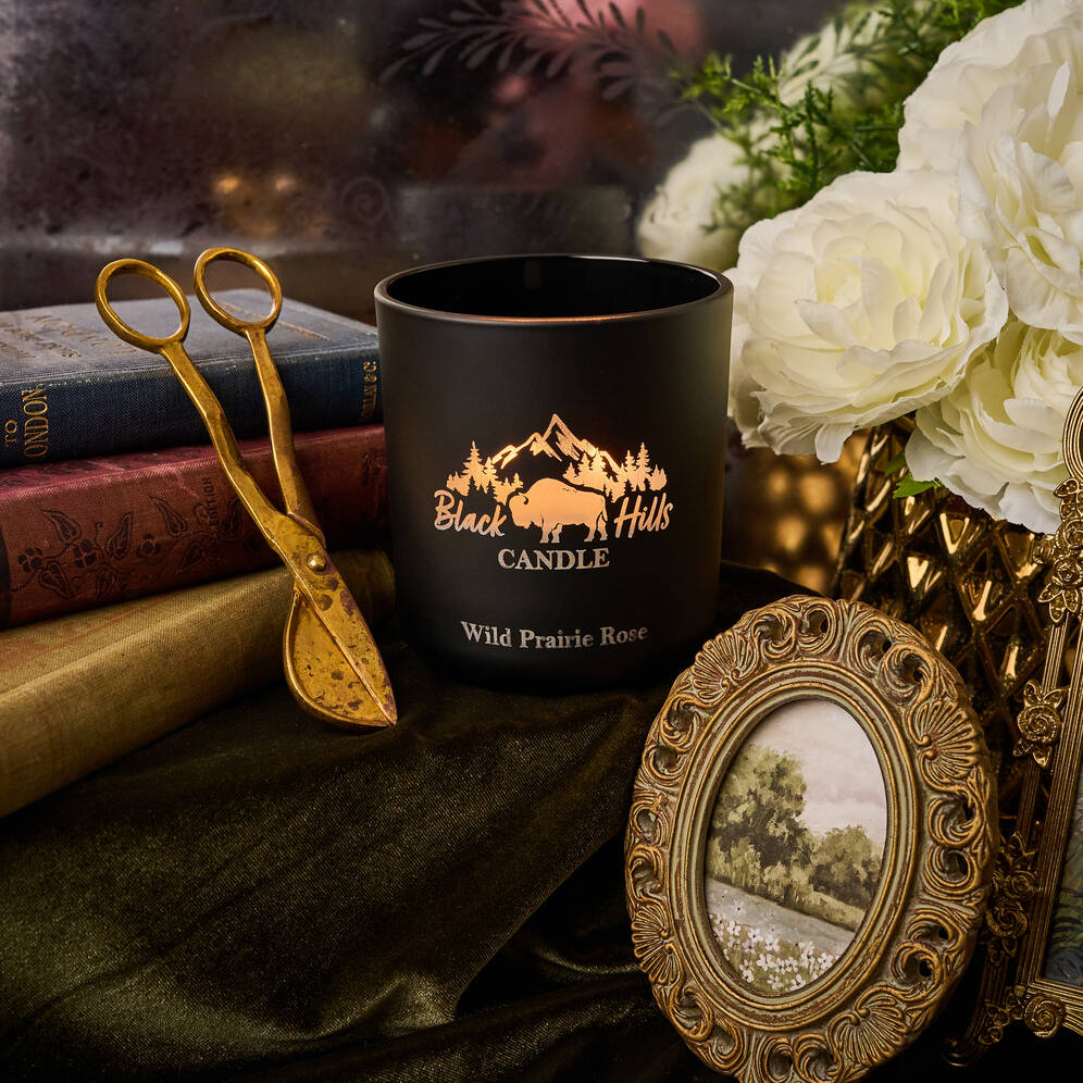 Black candle with 'Black Hills Candle' logo on a surface with decorative items like a gold frame and white flowers.