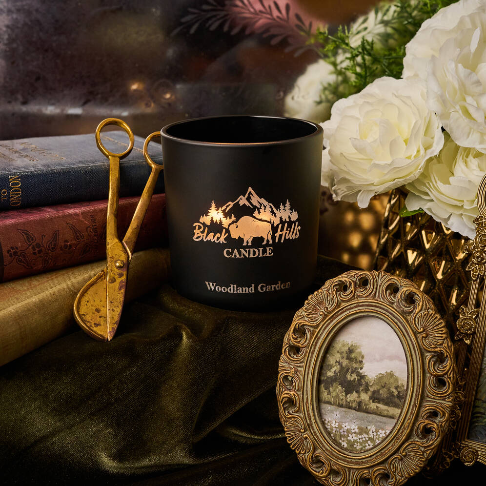 Black candle with 'Black Hills Candle' logo on a decorative surface with books and flowers.